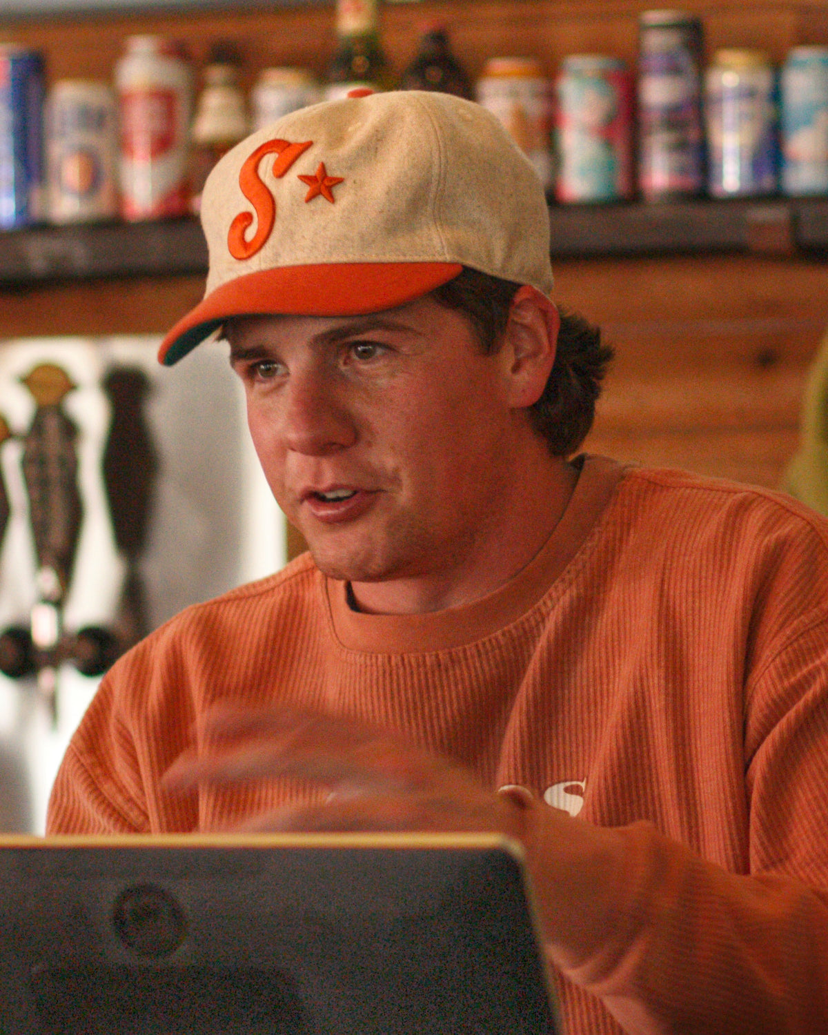 Man wearing a cap and orange sweater in a casual setting with shelves in the background