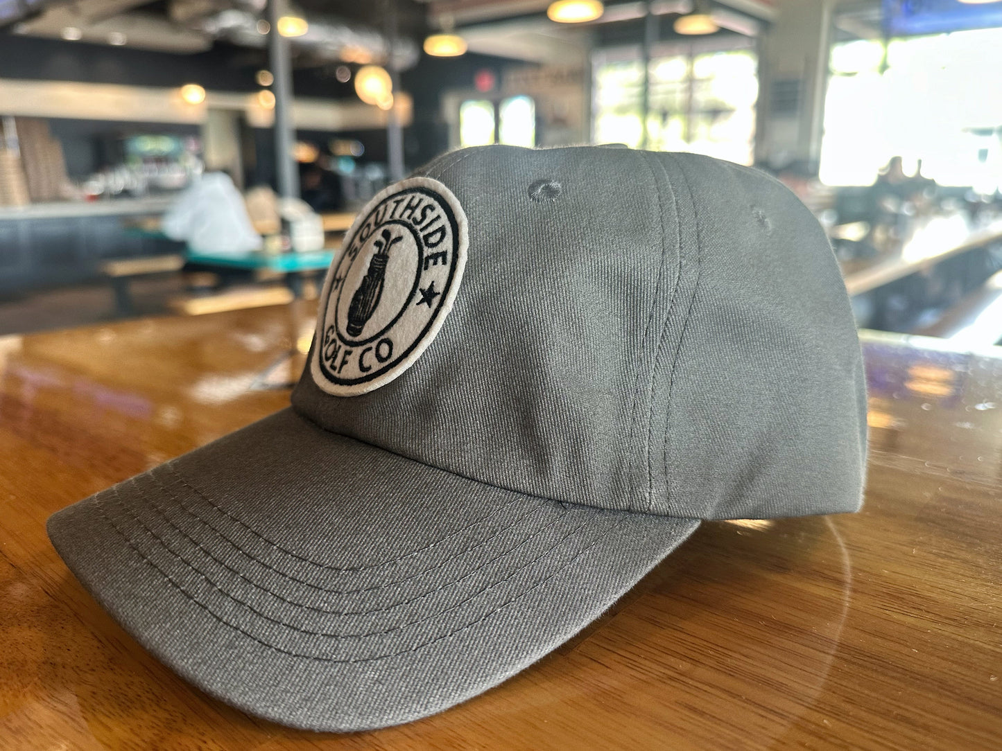 Southside Golf Co gray dad hat with embroidered logo patch, angled side view on wooden table indoors.