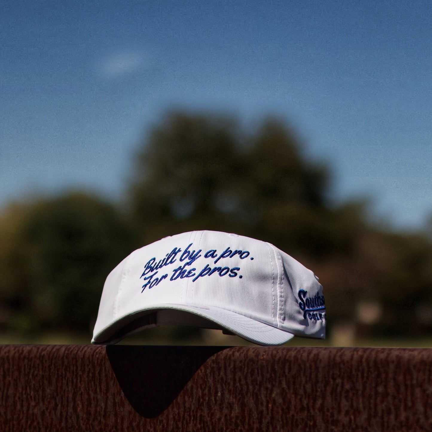 White lightweight performance rope golf hat with "Built by a pro. For the pros." embroidery resting on a steel railing at a golf course.