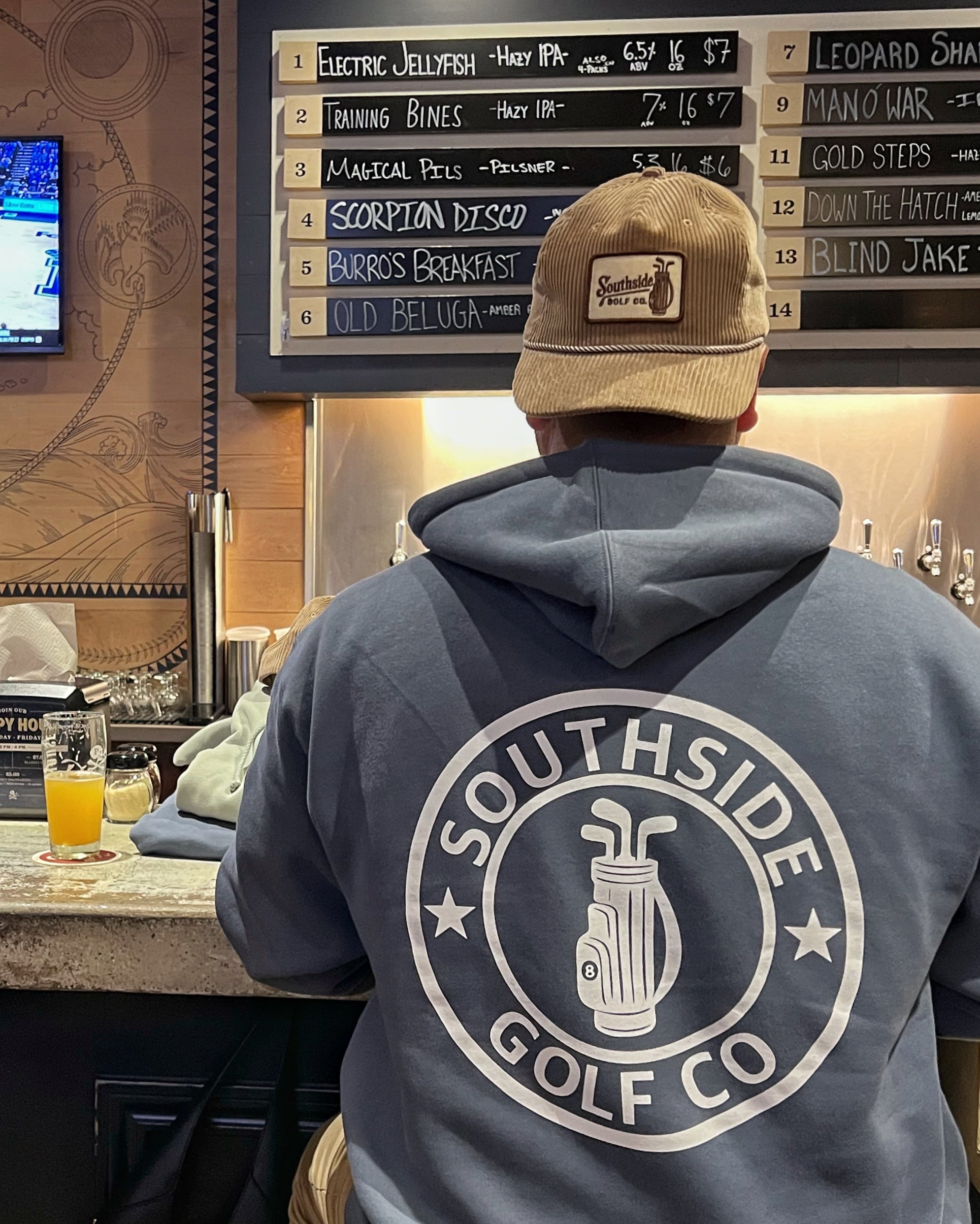 Back view of a person wearing a Southside Golf Co blue hoodie with large circular logo in a bar setting.
