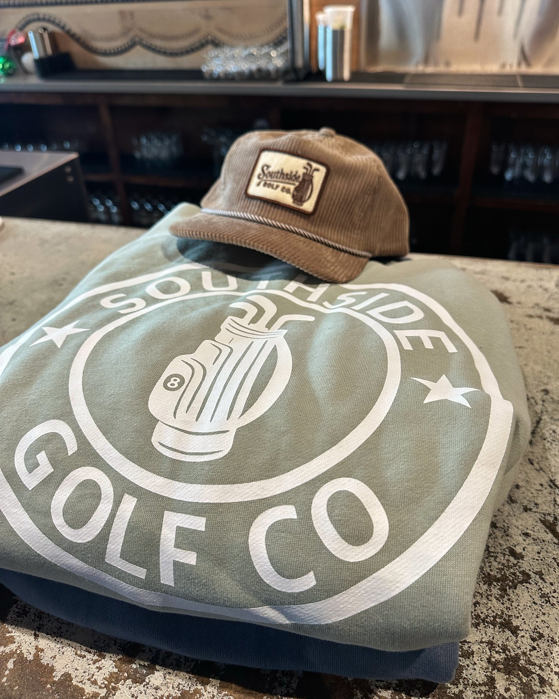 Southside Golf Co green hoodie with large back logo and corduroy golf hat on bar counter. 